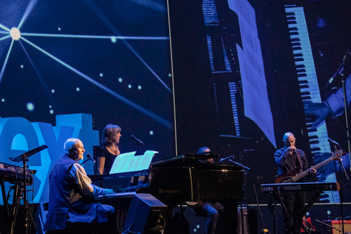 Three people on stage perform with a piano, keyboard, guitar, and vocals